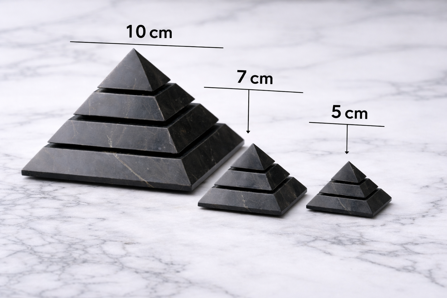 Three polished shungite pyramids in 10 cm, 7 cm, and 5 cm sizes displayed on a white marble surface, with size labels showing the different pyramid dimensions.