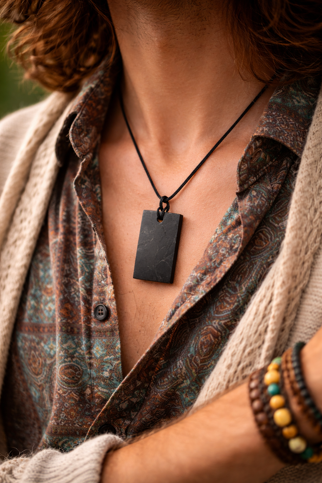 Rectangular black shungite pendant on a black cord worn by a bohemian male model from a side angle, shown on chest with patterned shirt and earthy layered clothing, face not visible.