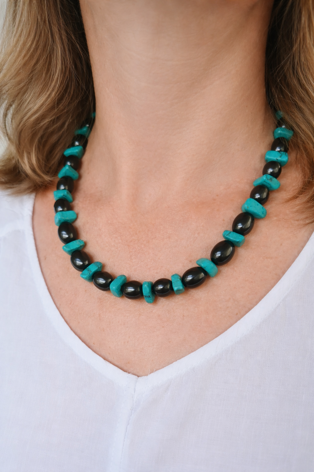 Shungite and raw turquoise beaded necklace worn on a woman’s neck with soft wavy hair and white blouse, close-up natural style jewelry photo