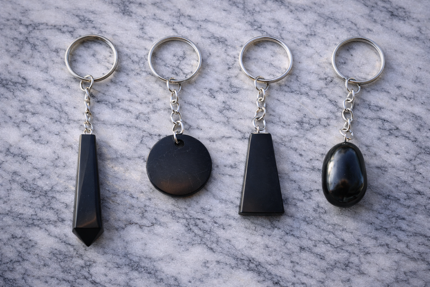 Four black shungite keychains in different shapes displayed side by side on a white marble surface with silver key rings.