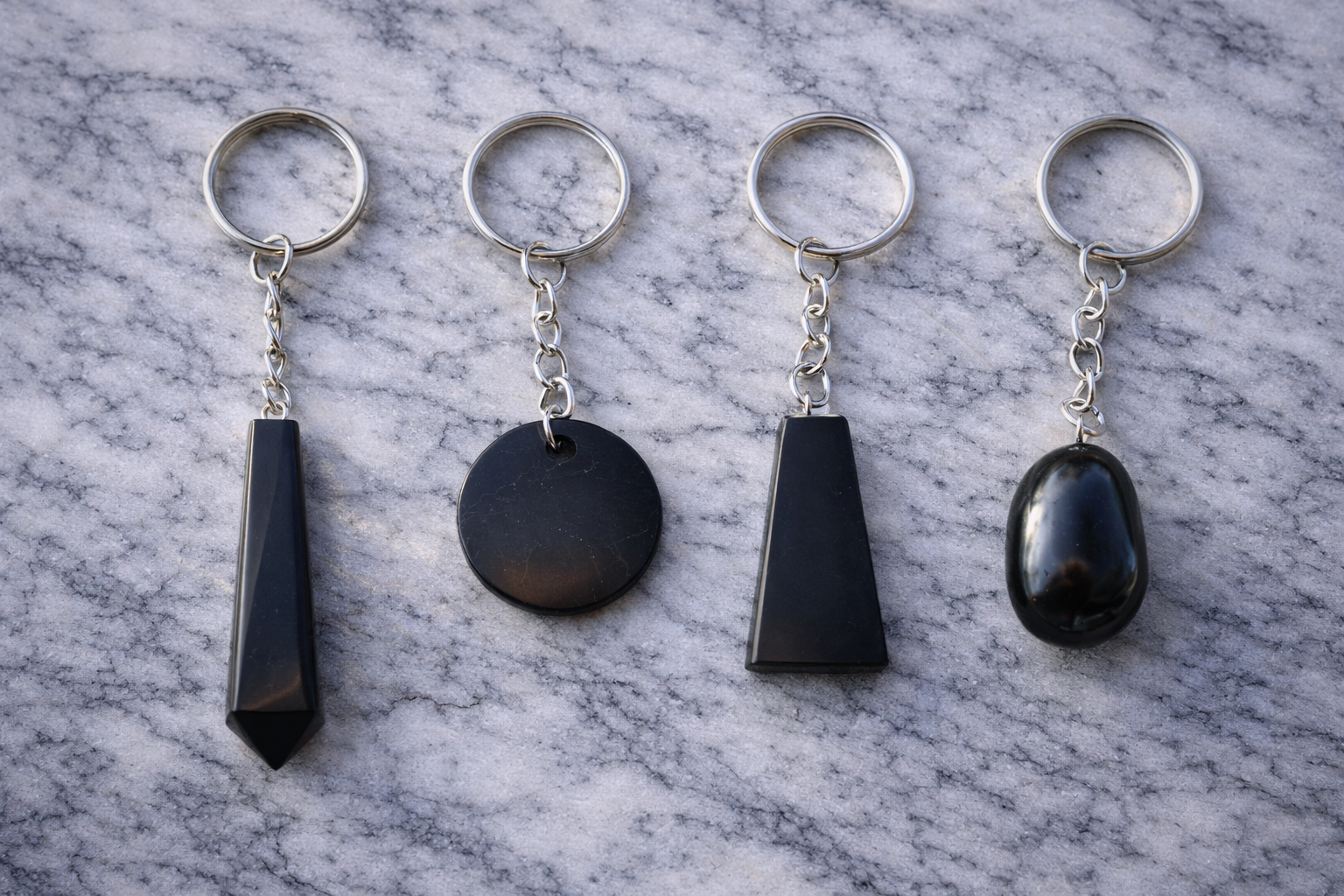 Four black shungite keychains in different shapes displayed side by side on a white marble surface with silver key rings.