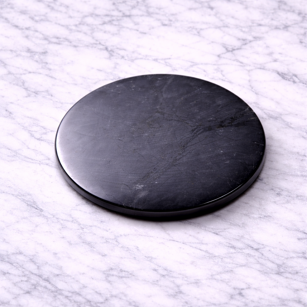 Polished black shungite EMF protection plate (50mm) resting on a white marble surface, showing natural stone veining and smooth glossy finish.