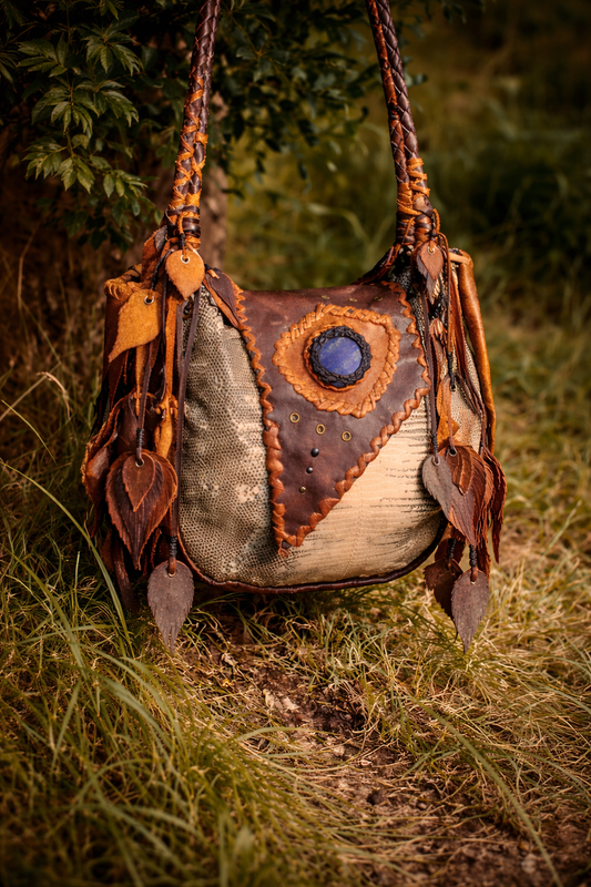 Handcrafted leather shoulder bag in earthy tones, featuring braided straps, leaf tassels, and a blue stone centerpiece, photographed outdoors among grass and greenery in soft natural light.