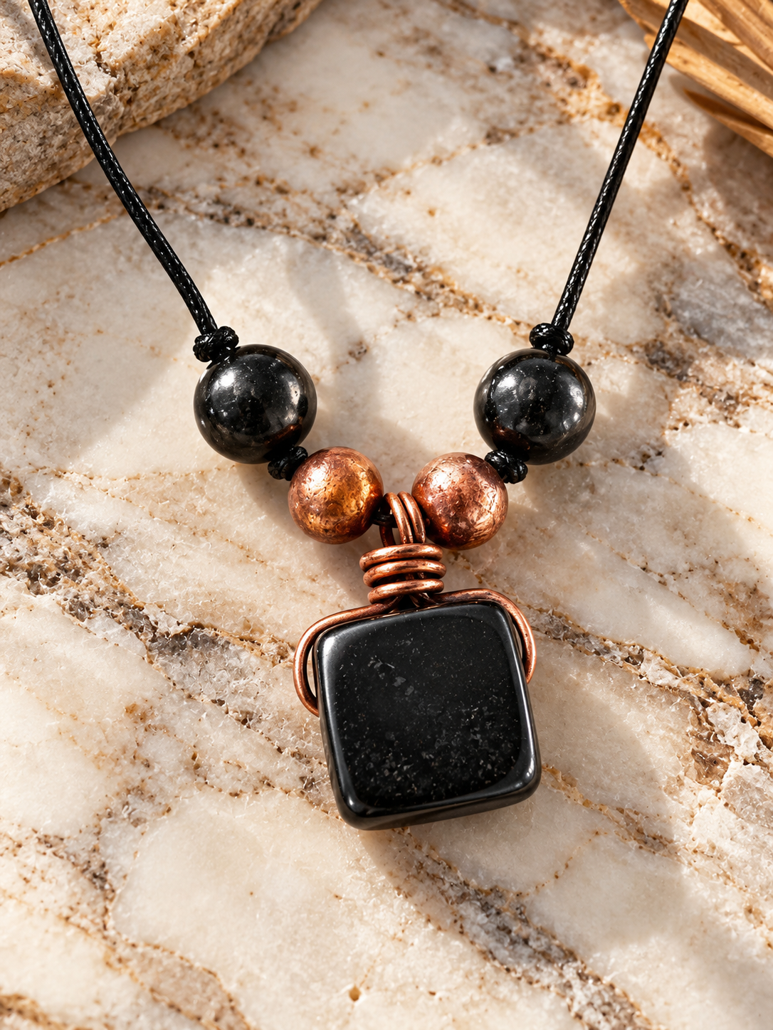 Handmade black shungite square pendant wrapped in copper wire, accented with copper and polished shungite beads on a black cord necklace, displayed on natural cream marble.