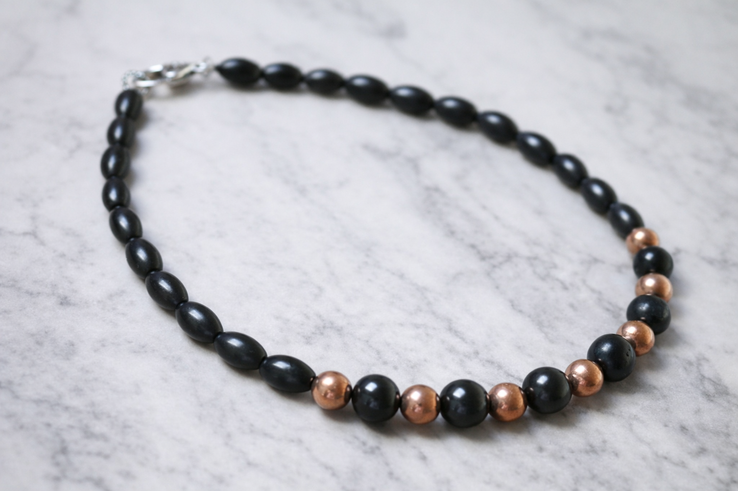 Handmade shungite and copper bead necklace arranged in a circle on a light marble surface with soft natural lighting and silver clasp visible