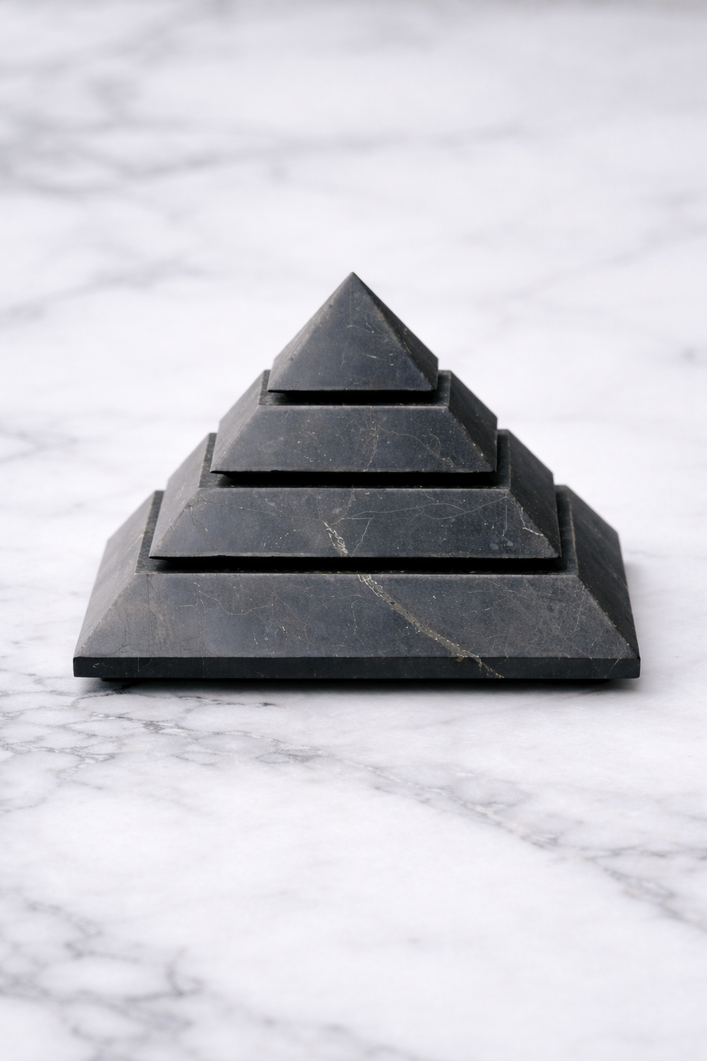 Front-facing polished shungite pyramid displayed on a white marble surface, showing stepped layers and natural stone veining in a minimalist product setting.