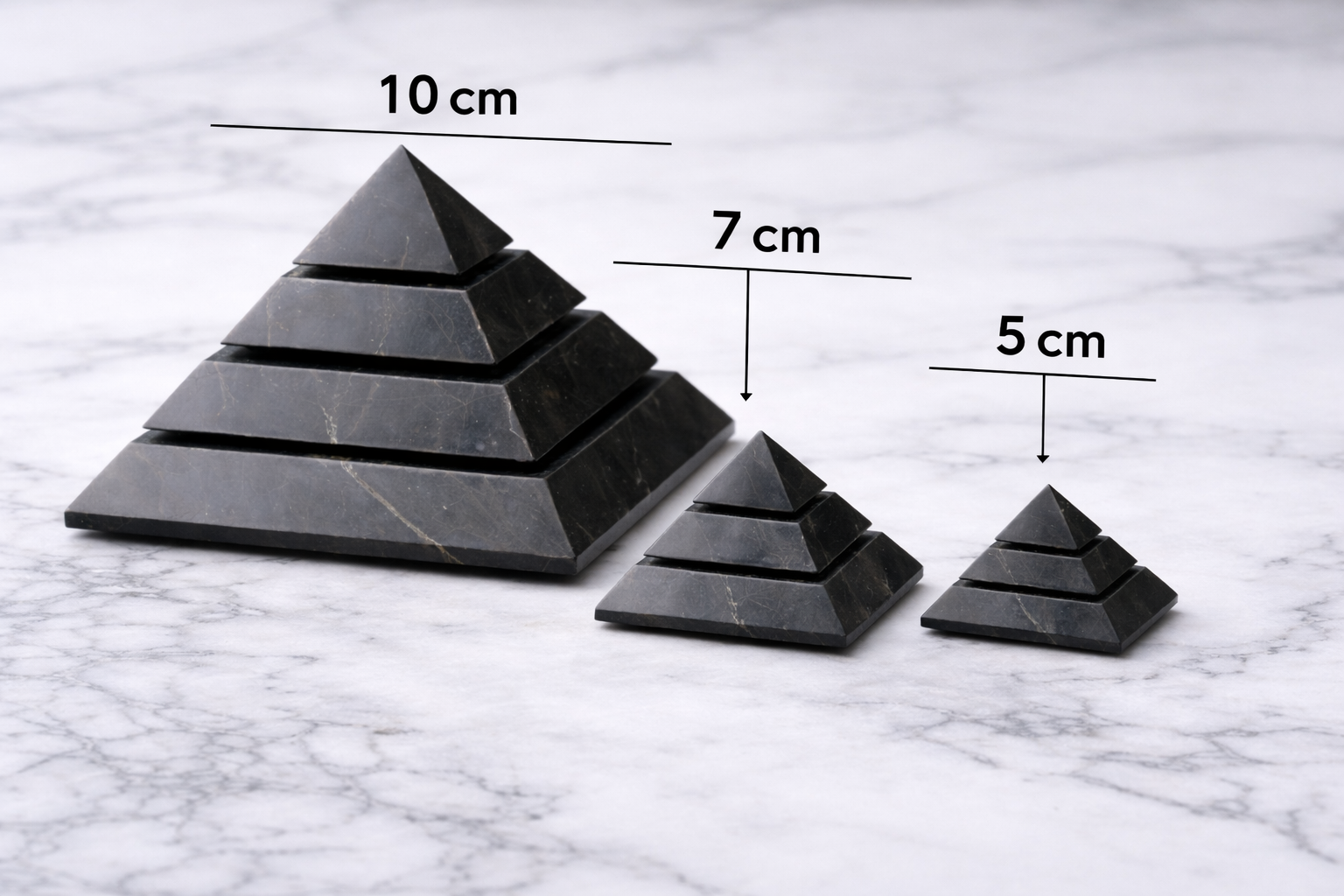Three polished shungite pyramids in 10 cm, 7 cm, and 5 cm sizes displayed on a white marble surface, with size labels showing the different pyramid dimensions.
