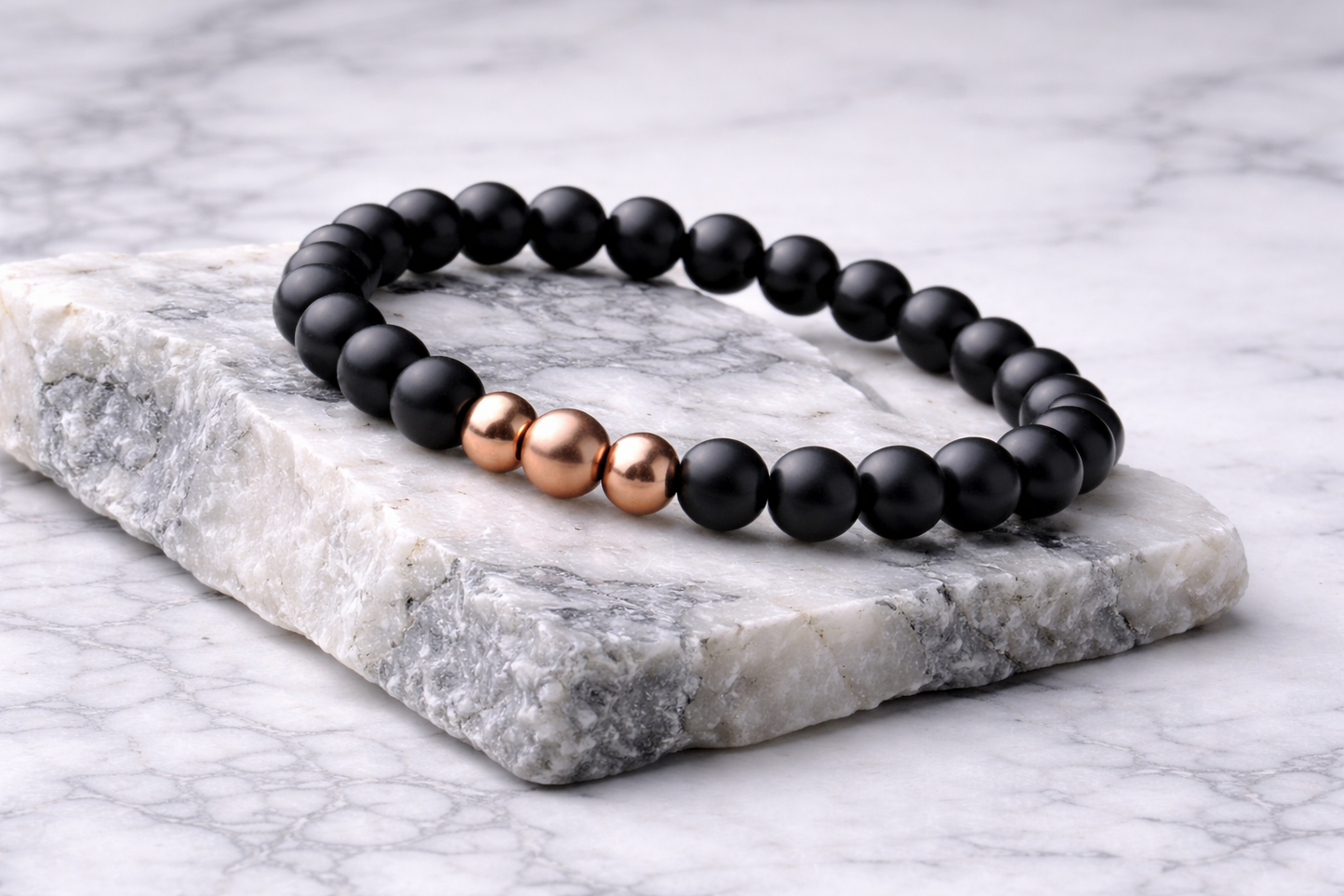 Black shungite beaded bracelet with three polished copper center beads resting on a white marble stone, photographed from a side angle with soft natural lighting.