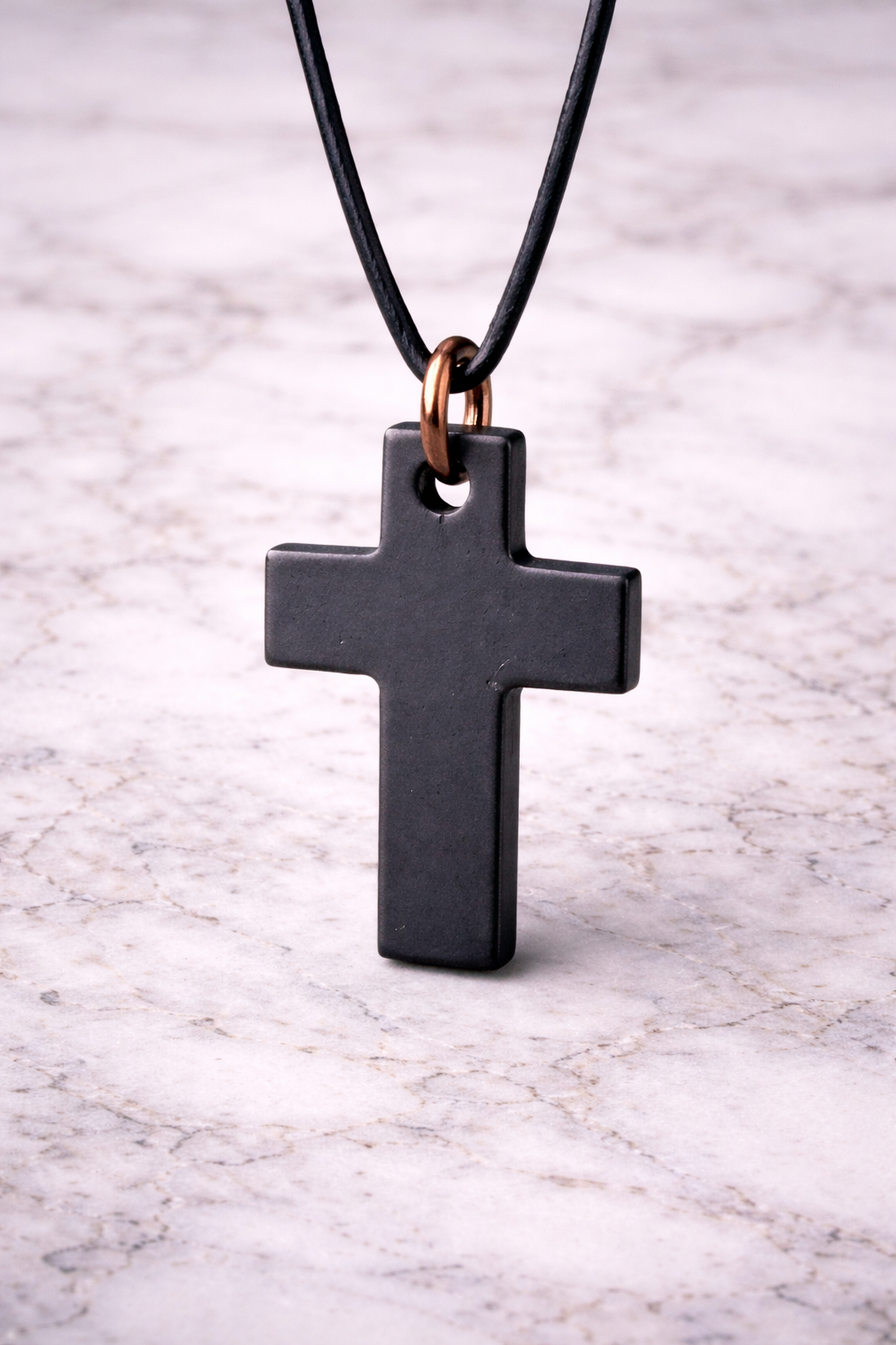 Black shungite cross pendant hanging on a black leather cord with copper hoop, photographed on a white marble background, showing smooth polished finish.