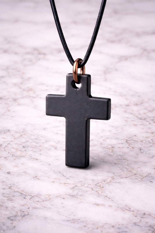 Black shungite cross pendant hanging on a black leather cord with copper hoop, photographed on a white marble background, showing smooth polished finish.