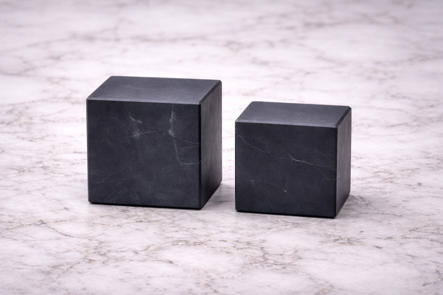 Two matte black shungite cubes in different sizes displayed side by side on a white marble surface, highlighting their smooth polished finish and natural stone texture.