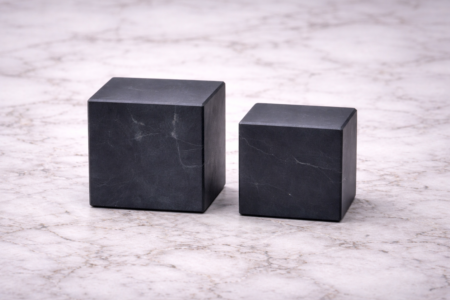 Two matte black shungite cubes in different sizes displayed side by side on a white marble surface, highlighting their smooth polished finish and natural stone texture.