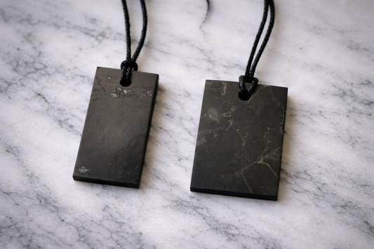 “Two rectangular shungite pendants on black cords resting on white marble surface”