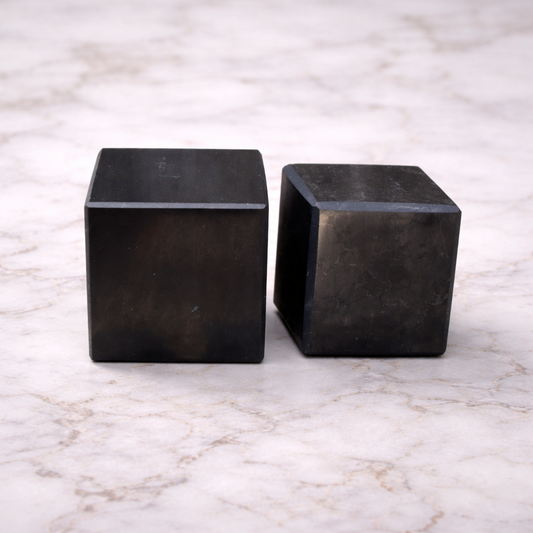 Two polished black shungite cubes displayed on a white marble surface, showcasing natural stone texture and minimalist crystal decor for grounding and energy balance.