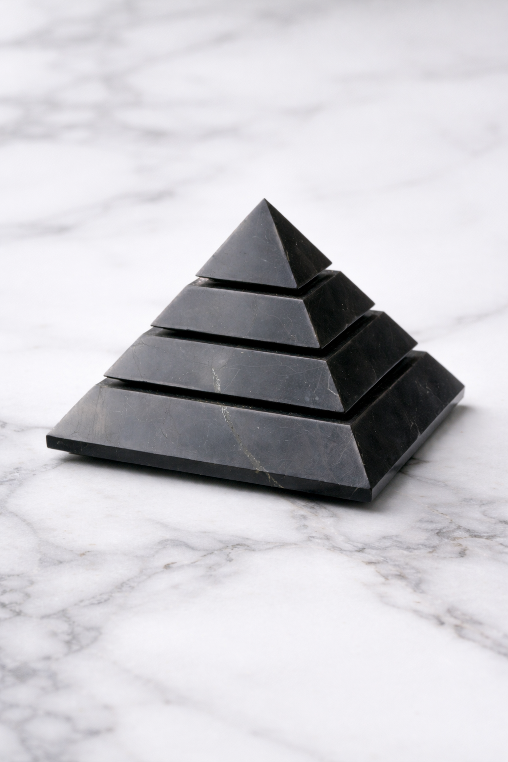  polished shungite pyramid displayed on a white marble surface, showing stepped layers and natural stone veining in a minimalist product settinngs