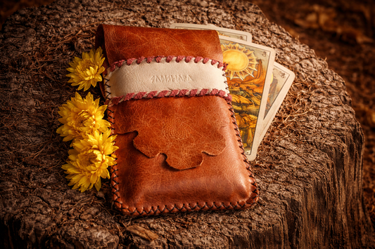 Handcrafted recycled leather tarot deck pouch resting on a tree stump, featuring a braided cream band, floral detail, and a tarot deck partially tucked inside, styled in warm natural light with yellow wildflowers for an earthy, ceremonial feel.