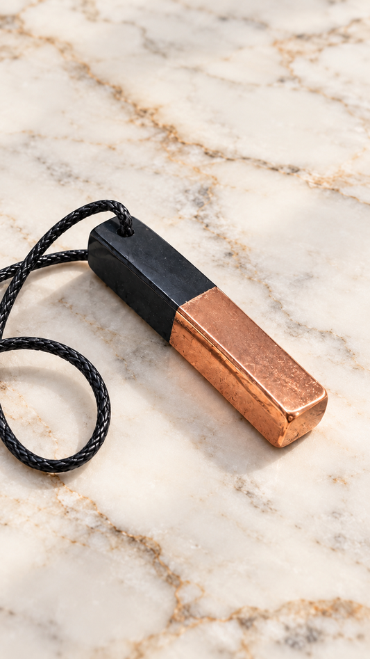 Rectangular black shungite pendant with a polished copper-coated half and braided black cord, resting on a natural white marble surface with soft lighting.