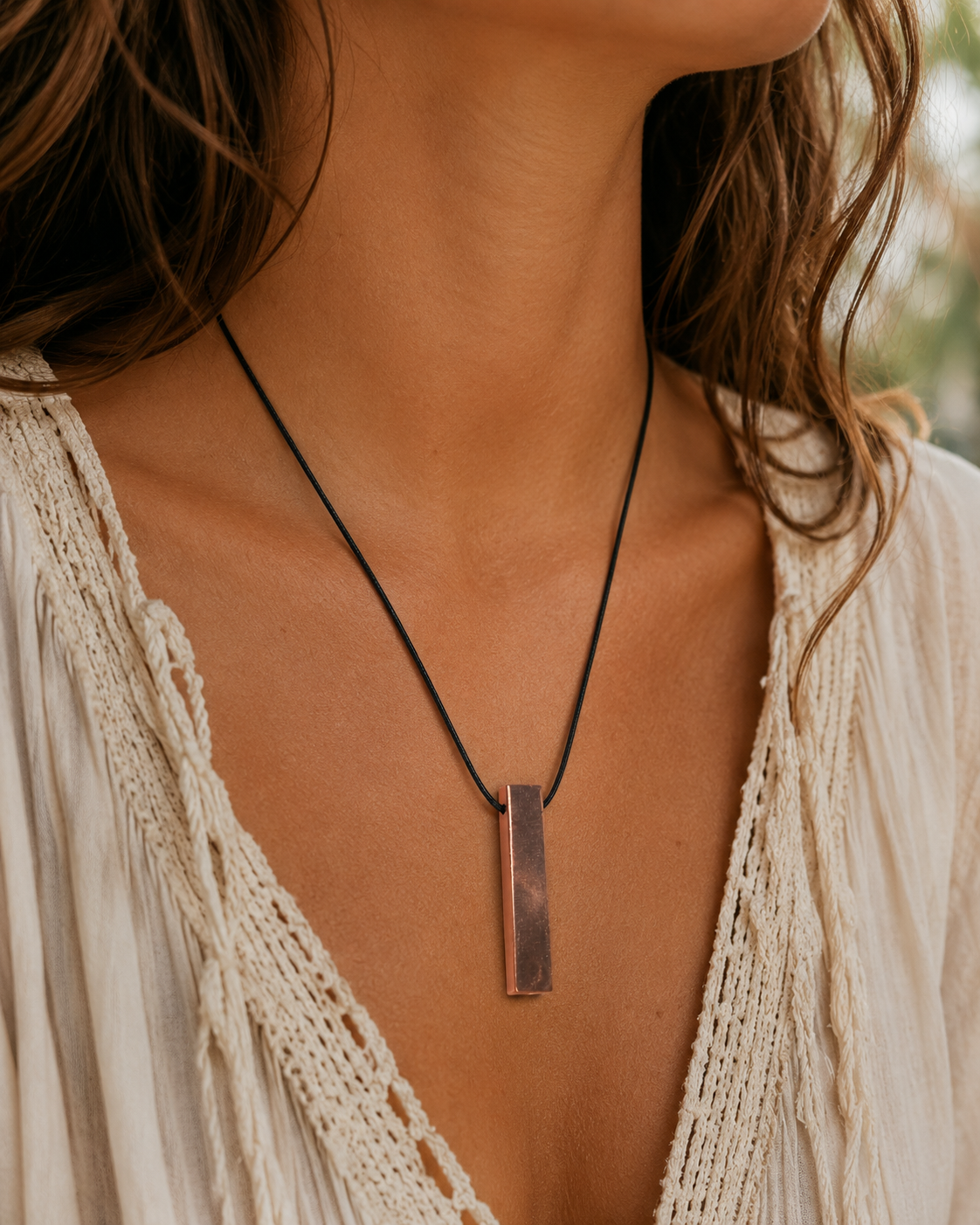 Boho woman wearing a handmade copper-coated shungite bar pendant necklace on a black cord, styled with a soft cream bohemian blouse in warm natural lighting.
