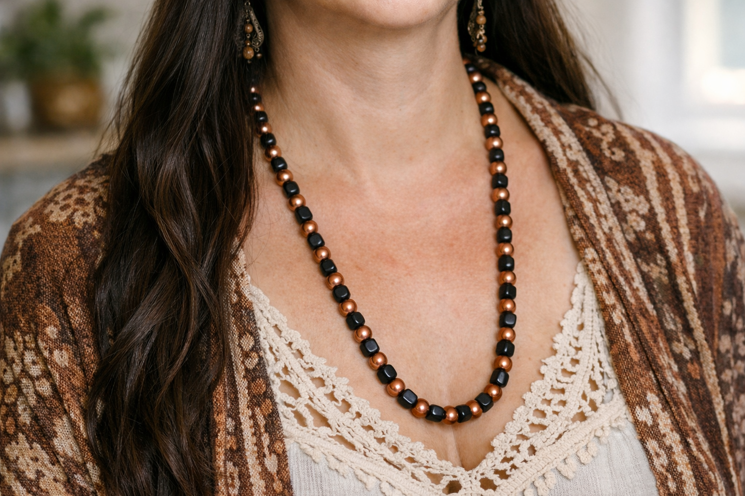 Close-up photograph of a woman wearing a handmade beaded necklace with alternating black shungite cube beads and copper-coated round beads. She has long dark hair and a soft bohemian look, styled in a cream lace crochet top with a patterned shawl, set against a warm, softly blurred natural background.
