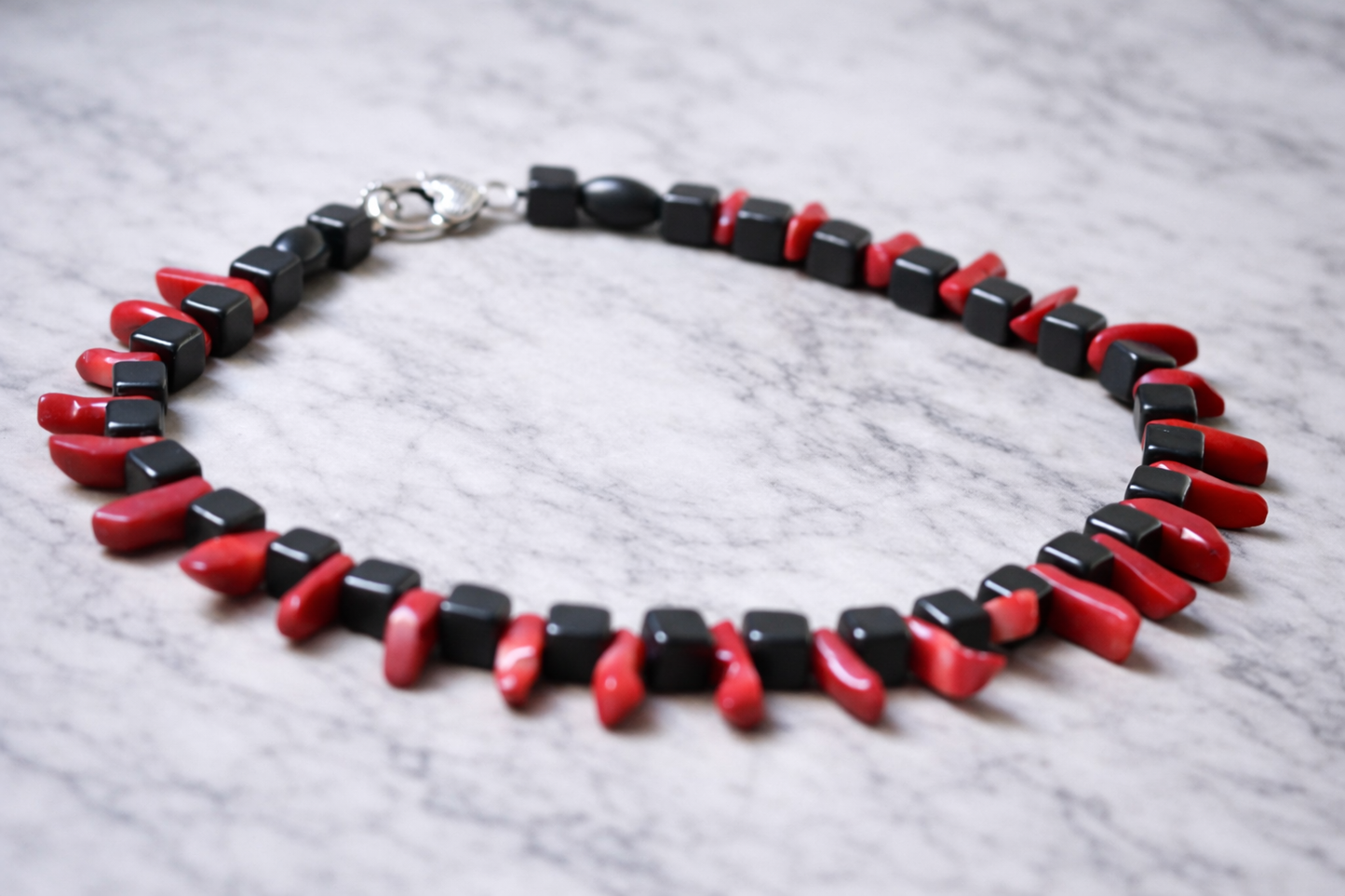 Handmade shungite cube bead necklace with red coral accents arranged in a circle on a light marble surface with soft natural lighting