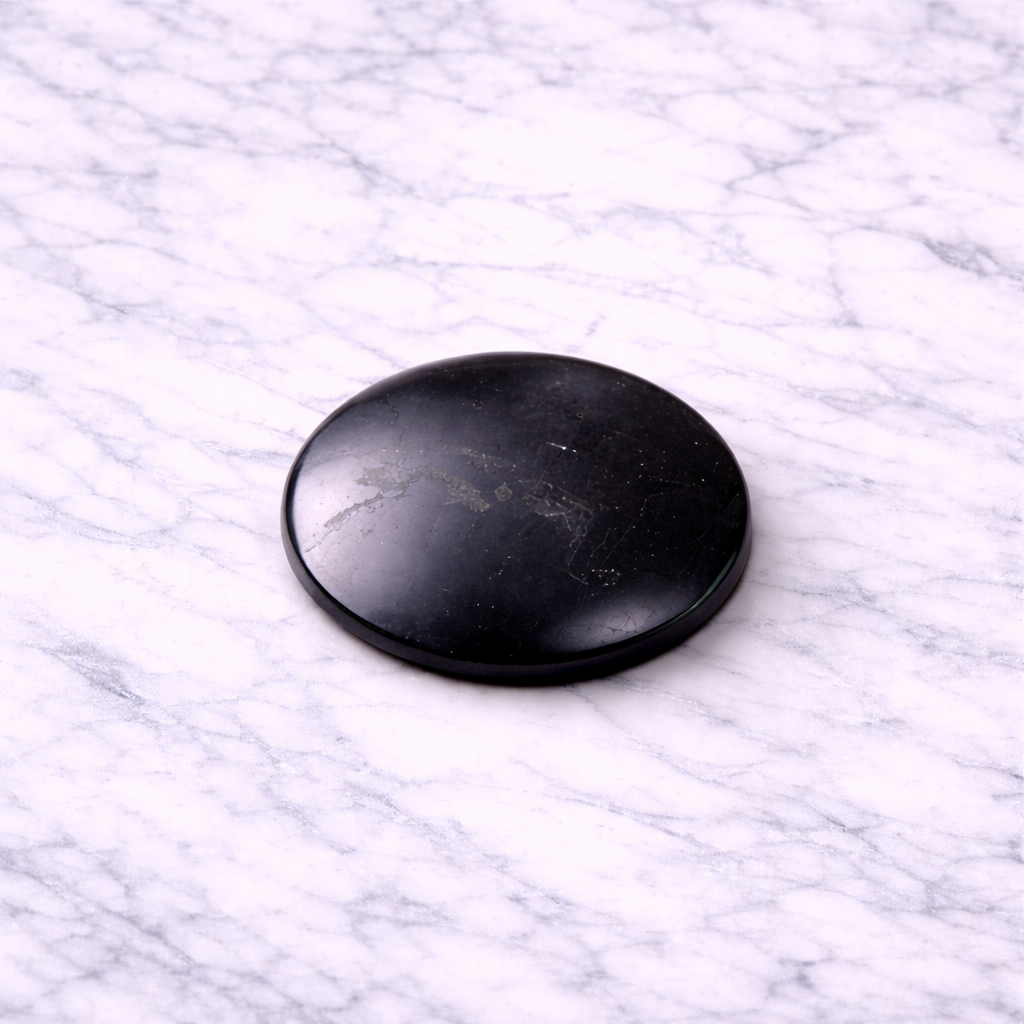 Polished black shungite EMF protection plate convex (30mm) resting on a white marble surface, showing natural stone veining and smooth glossy finish.
