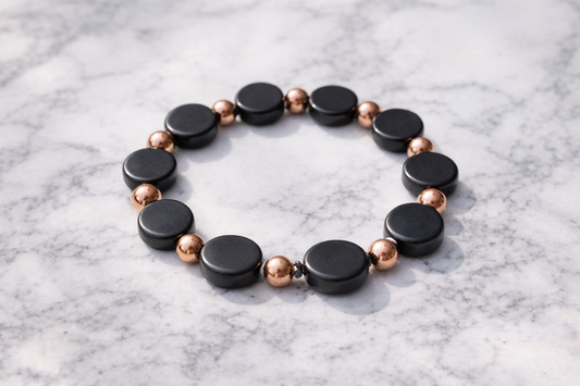 Black shungite and copper bead bracelet arranged in a circle on a white marble surface, with alternating matte stone and polished metal beads.
