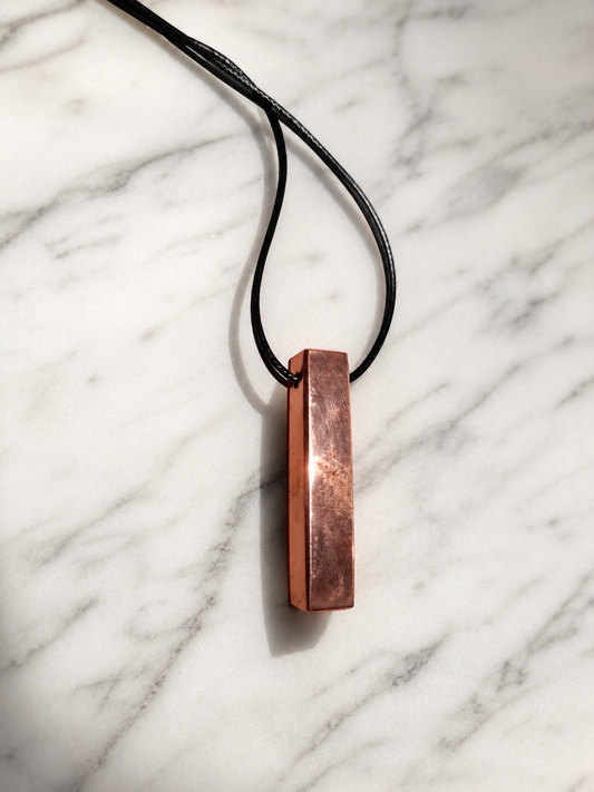 Handcrafted copper-coated shungite bar pendant necklace displayed on a white marble surface with soft natural lighting and gentle shadows, featuring a minimalist artisan design on a black cord.