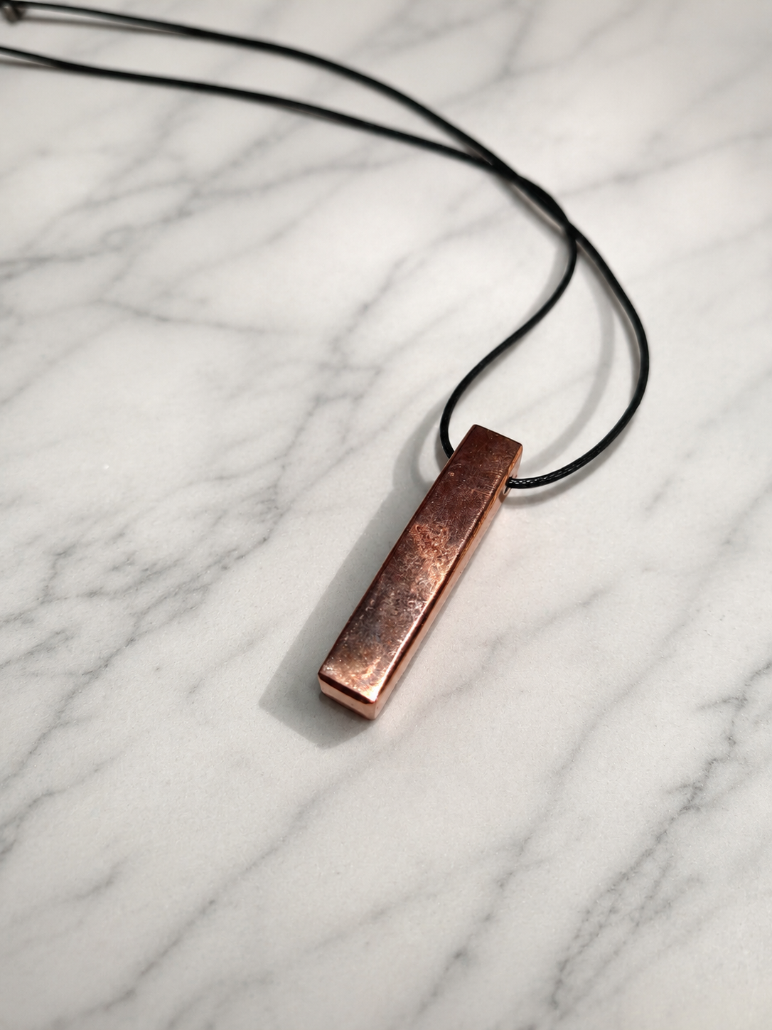 Handcrafted copper-coated shungite bar pendant necklace displayed on a white marble surface with soft natural lighting and elegant shadows, featuring a minimalist artisan design with a black cord.