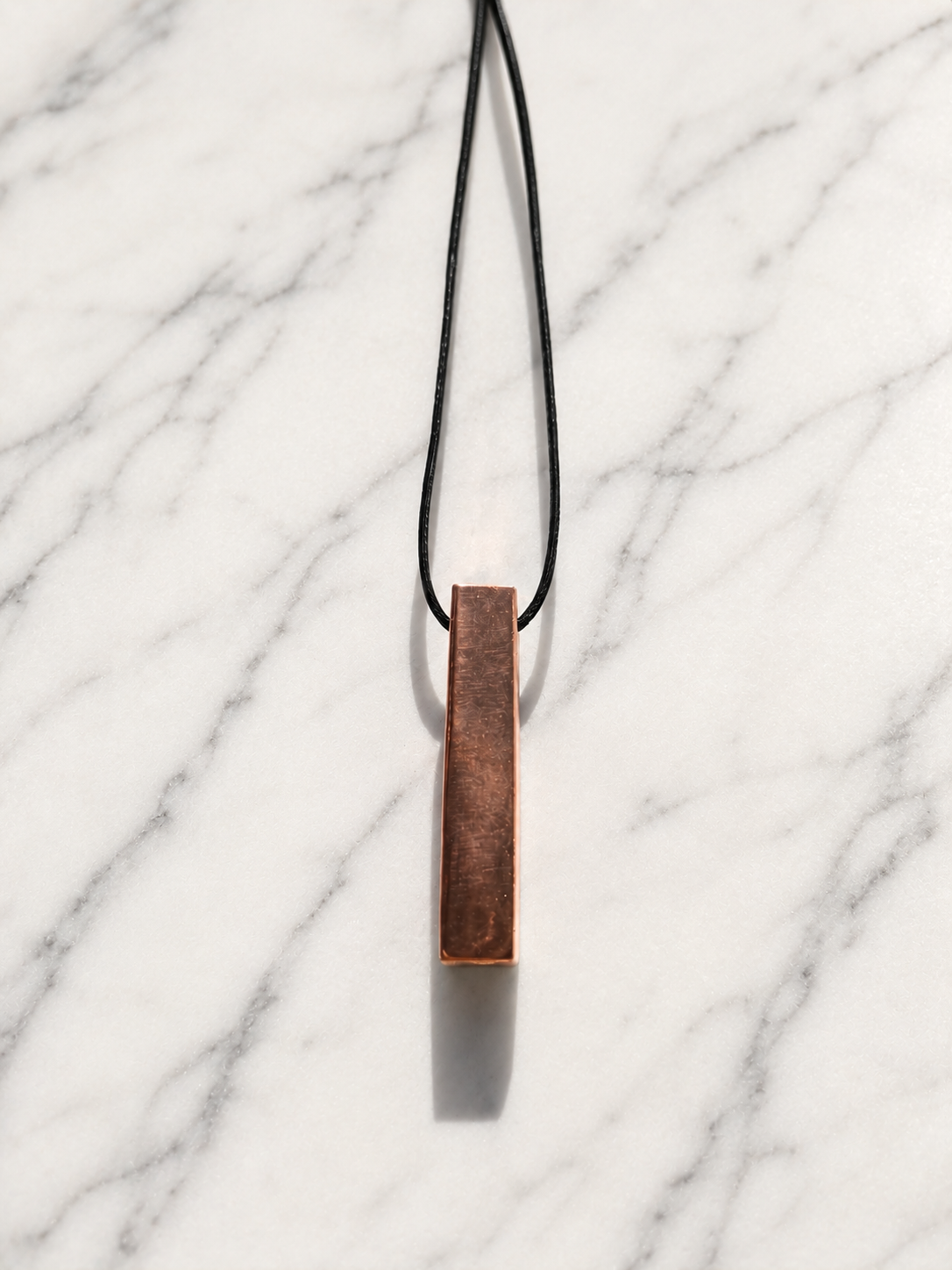 Handcrafted copper-coated shungite bar pendant necklace displayed on a white marble background with soft natural lighting and elegant shadows, featuring a minimalist artisan design on a black cord.