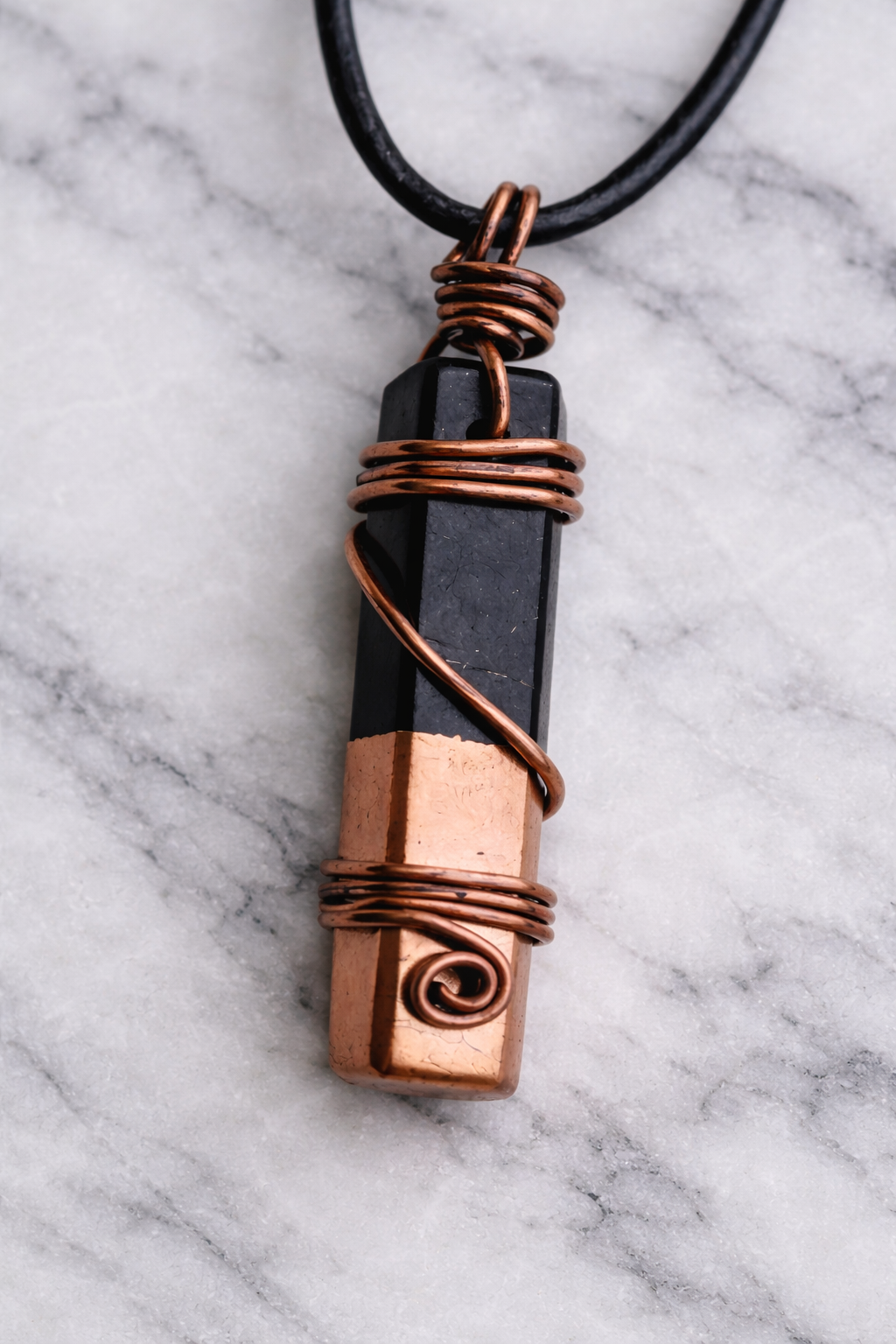 Close-up of a handmade shungite and copper wire-wrapped pendant on a black cord, resting on a white marble surface.