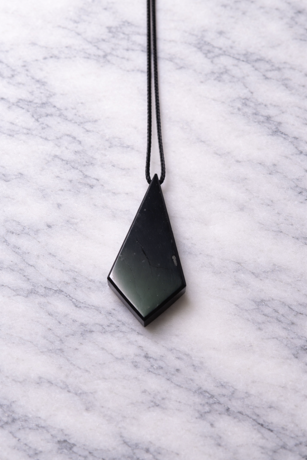 Polished black shungite pendant on a straight black cord, displayed on a white marble surface with soft natural lighting, highlighting its geometric diamond shape and subtle natural textures.