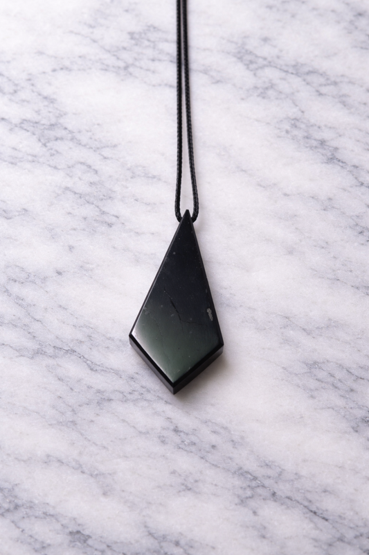 Polished black shungite pendant on a straight black cord, displayed on a white marble surface with soft natural lighting, highlighting its geometric diamond shape and subtle natural textures.