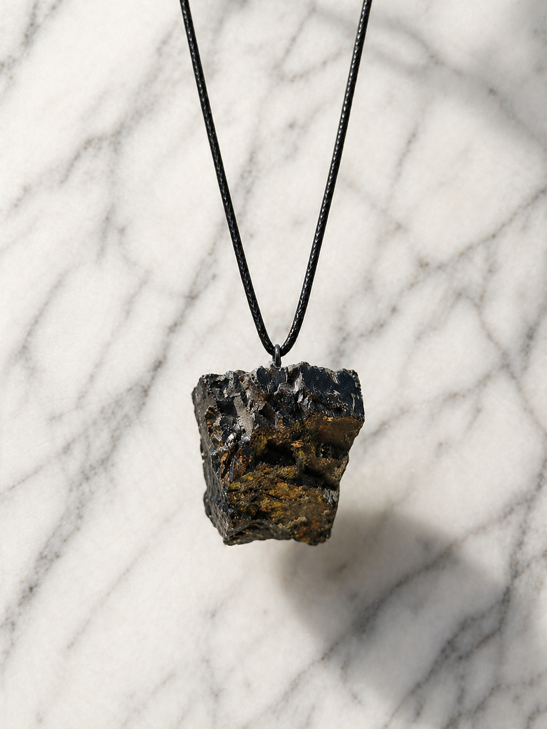 Raw shungite pendant hanging on black cord against white marble background with soft natural light