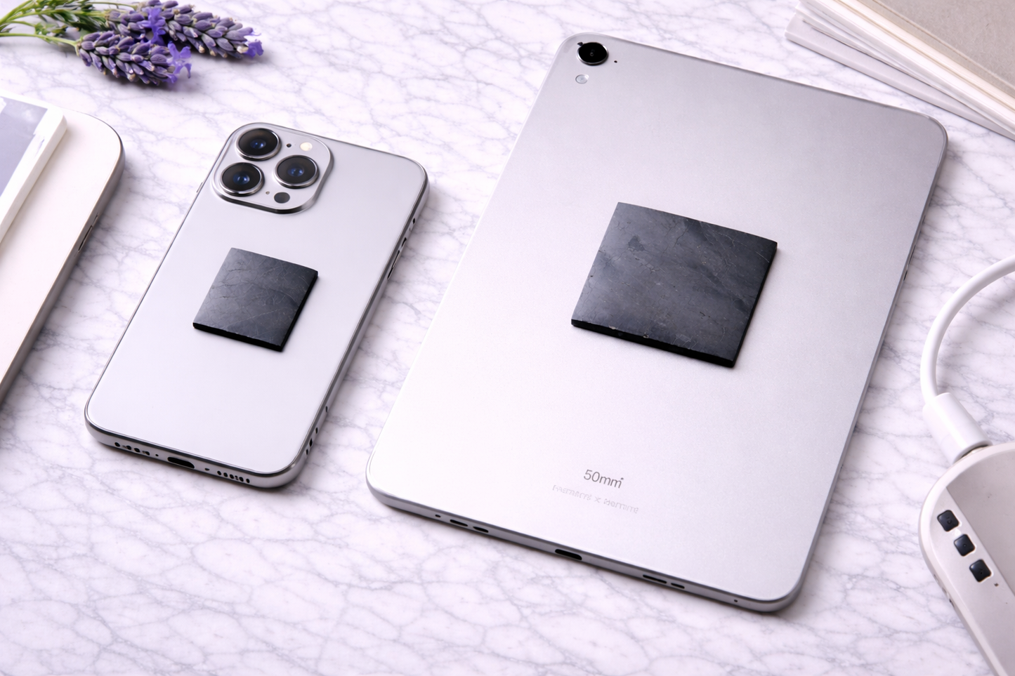 Silver smartphone and tablet lying on a white marble surface, each with a square black shungite EMF protection plate attached to the back, showing size difference between the phone plate and larger tablet plate.