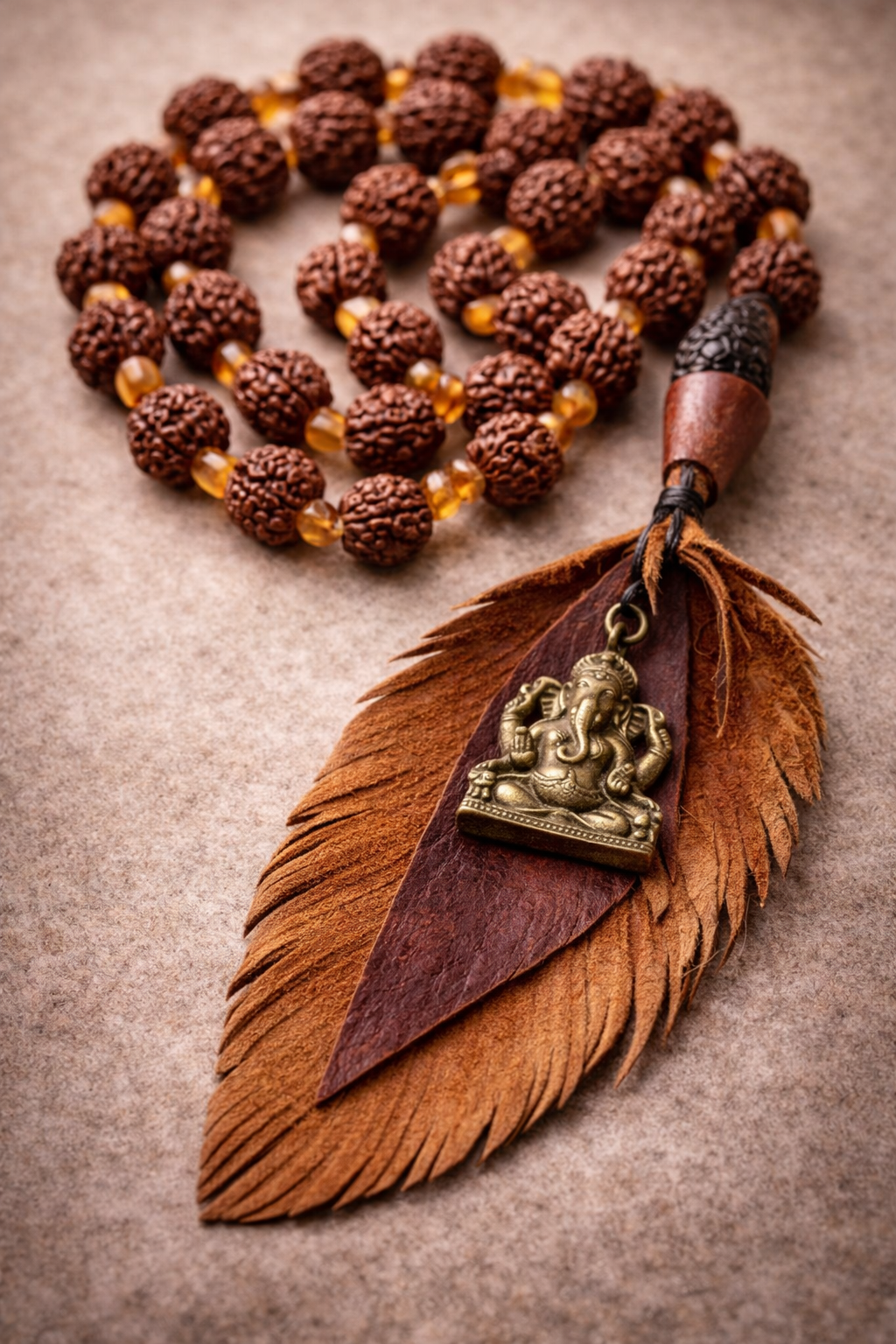 Close-up of a handmade Rudraksha mala with warm amber-toned spacer beads, finished with a layered recycled leather feather and a small brass Ganesh charm, resting on a soft neutral stone surface.