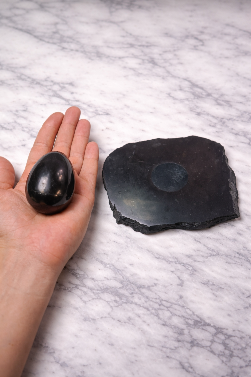 Polished shungite egg held in hand for size reference beside raw shungite base on white marble surface, showing glossy finish, natural texture, and approximate scale