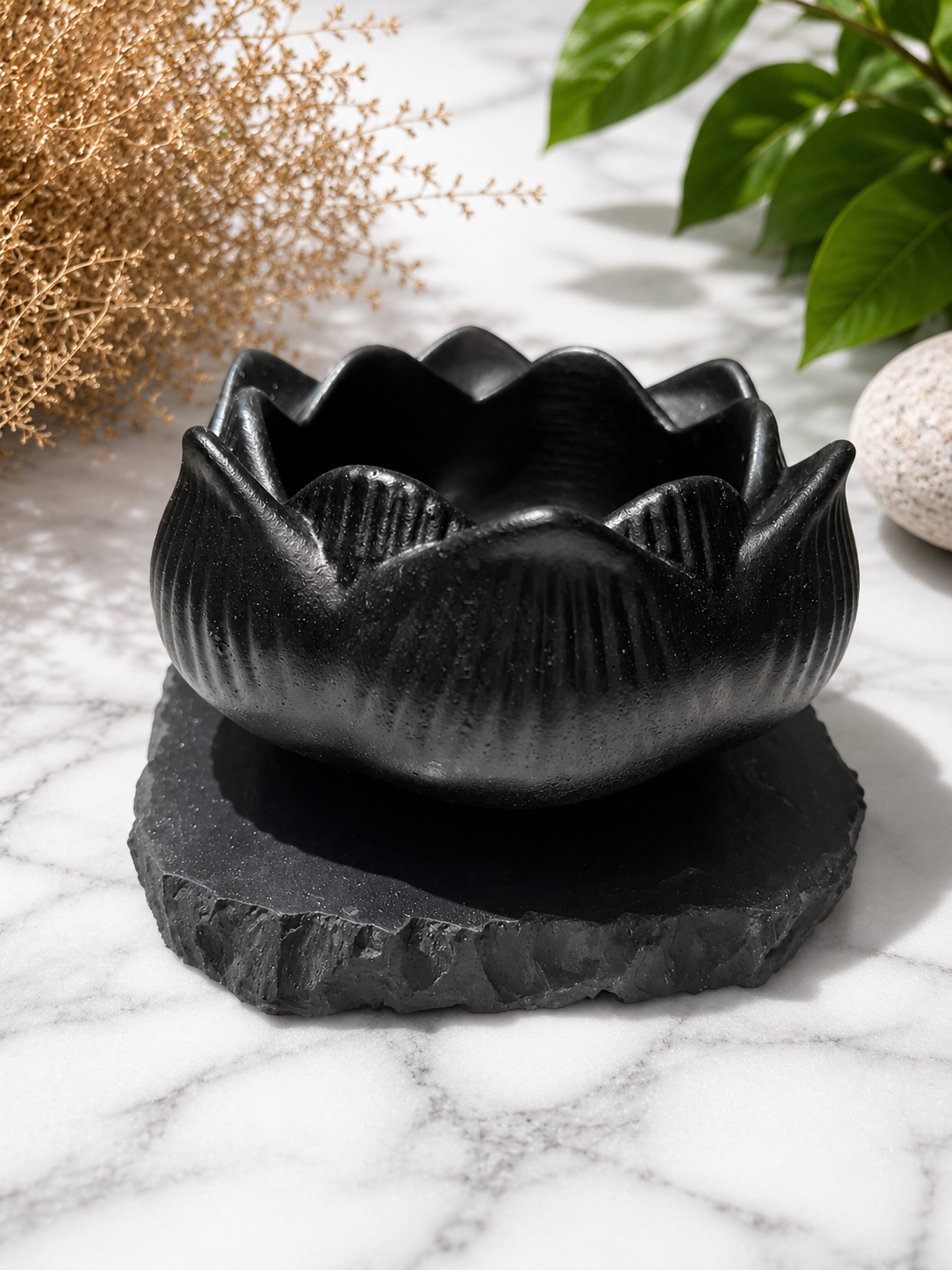 Black lotus-shaped shungite candle holder on a raw stone base, displayed on a marble surface with soft natural lighting, gentle shadows, and calming botanical decor in a minimalist spa-inspired setting.