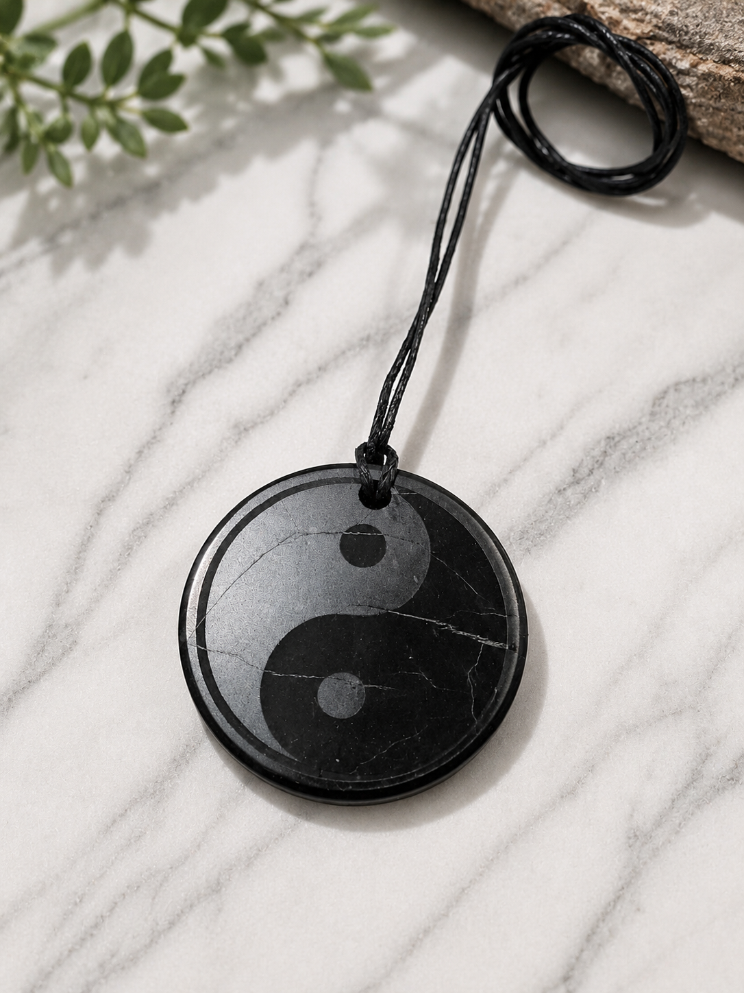 Round black shungite yin-yang pendant necklace handcrafted from genuine shungite stone, displayed on a white marble surface with soft natural lighting. The polished pendant features natural mineral veining and hangs from a black adjustable cord, creating a balanced and minimalist design inspired by the yin-yang symbol.