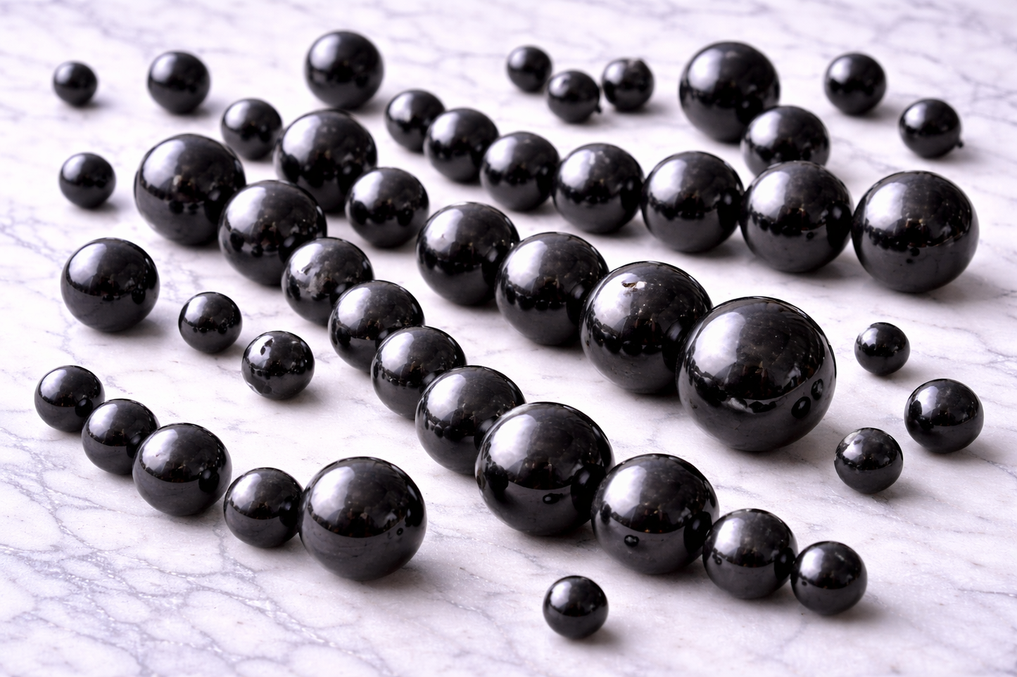 Loose polished black shungite beads in mixed sizes (approximately 6mm–12mm) scattered across a white marble surface, showing glossy reflective finish and natural variations, suitable for jewelry making and craft projects.