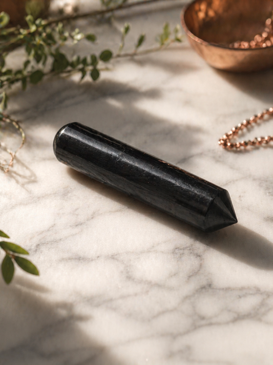 Close-up of a polished black shungite massage stick placed diagonally on a light marble surface, with soft natural lighting, gentle shadows, and subtle greenery and copper accents in the background.