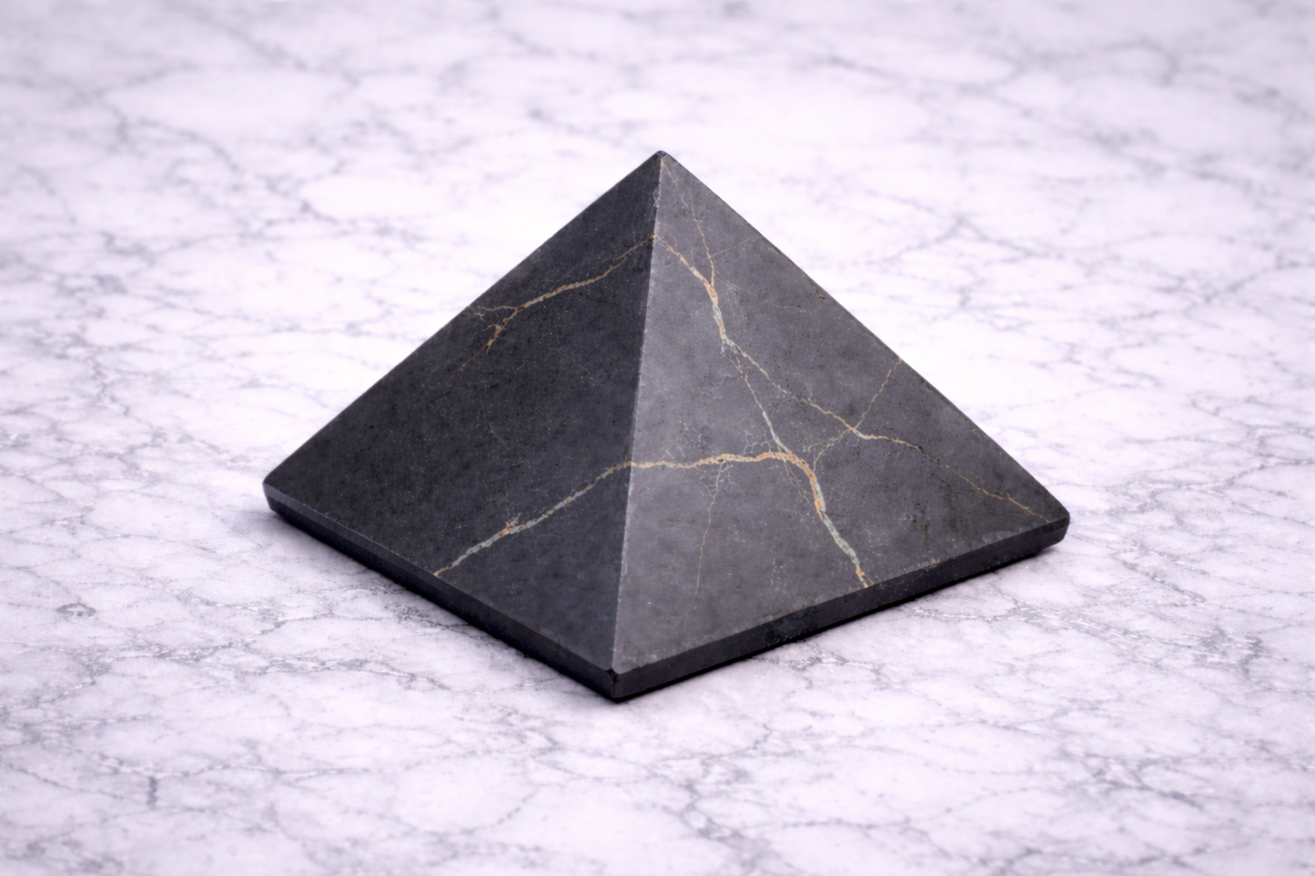 Matte black shungite pyramid with subtle natural veining displayed on a white marble surface, photographed from a front angle to show its geometric shape and smooth finish.
