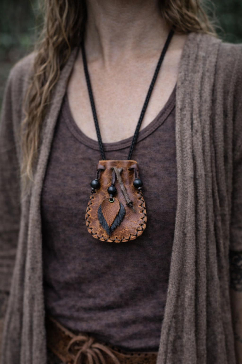 “Close-up of a woman from the neck down wearing a small handmade brown leather medicine bag on a black cord, photographed outdoors with a soft forest background.”