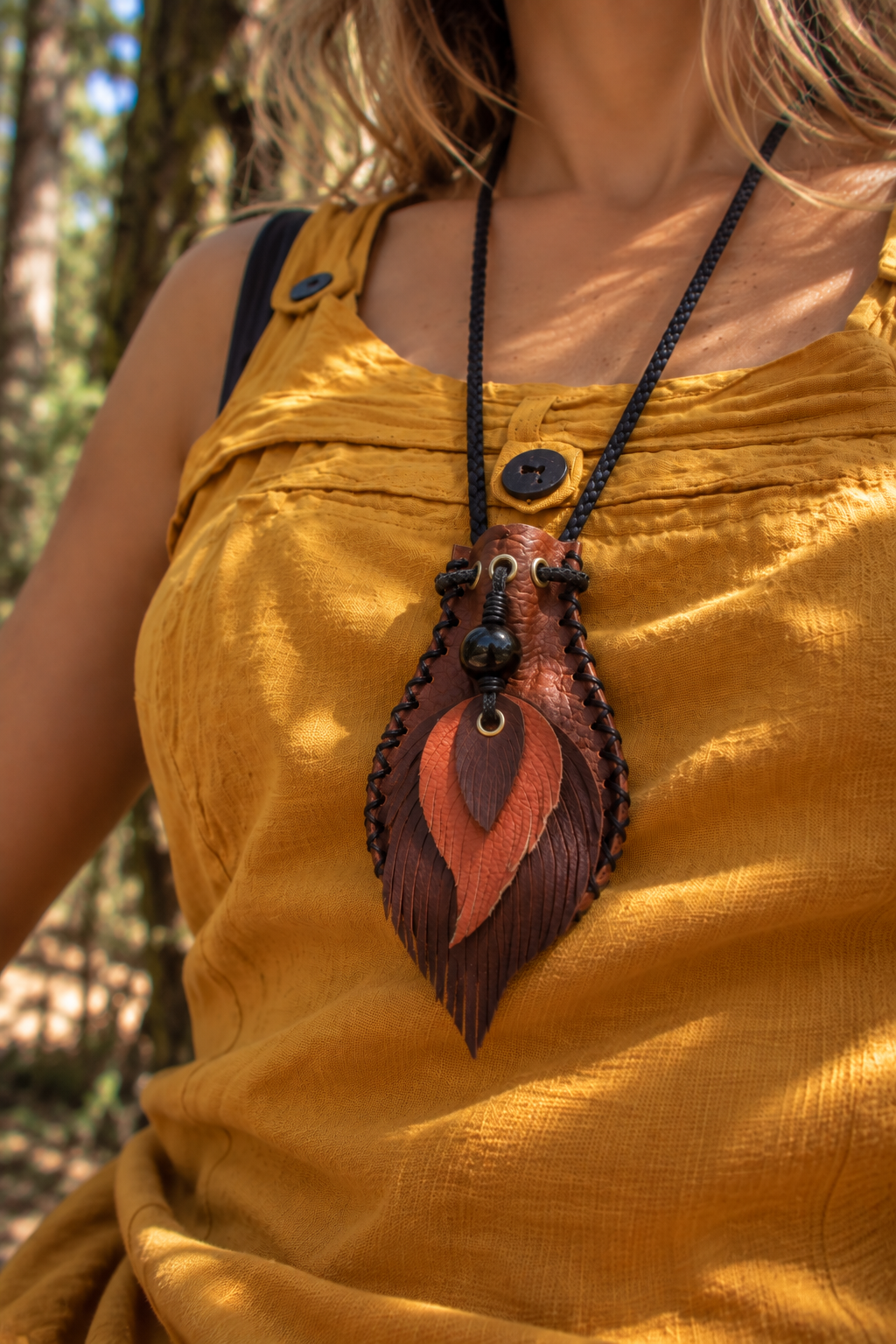 Handcrafted leather shungite pouch necklace with decorative leather feathers and a polished shungite bead, worn outdoors in a natural forest setting.