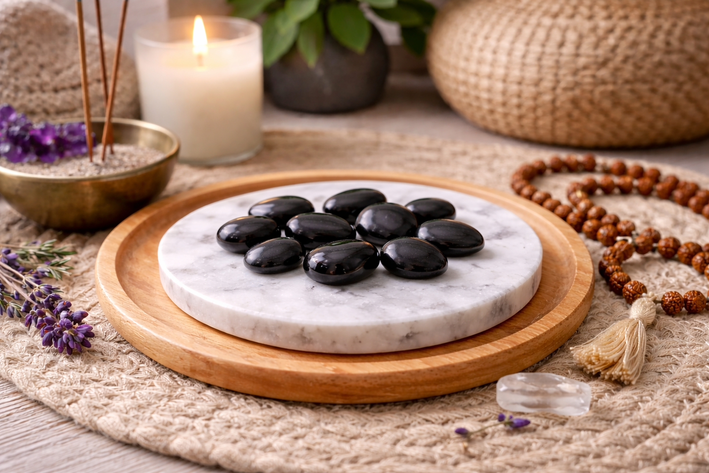 Small polished black shungite tumble stones arranged on a round white marble plate in a peaceful meditation setting, surrounded by a lit candle, incense bowl, lavender sprigs, rudraksha beads, and crystals on a natural woven mat.