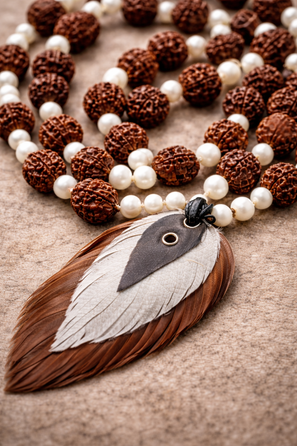 Handcrafted Rudraksha and pearl mala necklace arranged on a neutral stone surface, featuring textured brown Rudraksha beads, white pearls, and a layered feather pendant centerpiece.