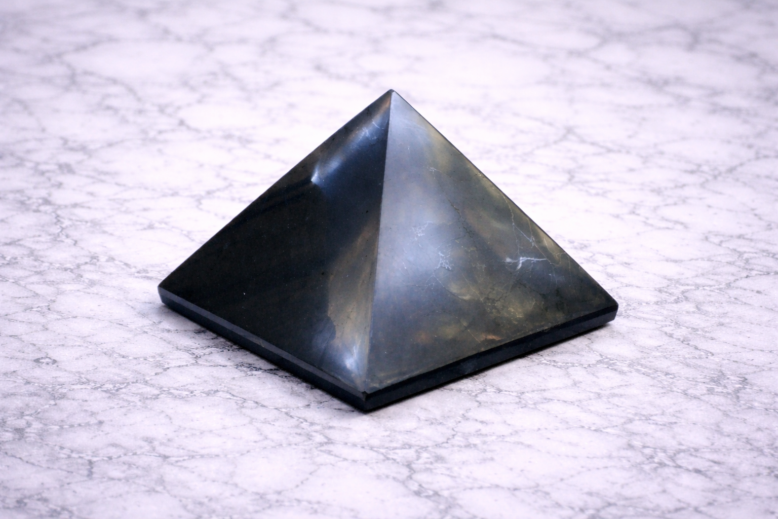 shiny black shungite pyramid displayed on a white marble surface, photographed from a front angle to show its geometric shape and smooth finish.