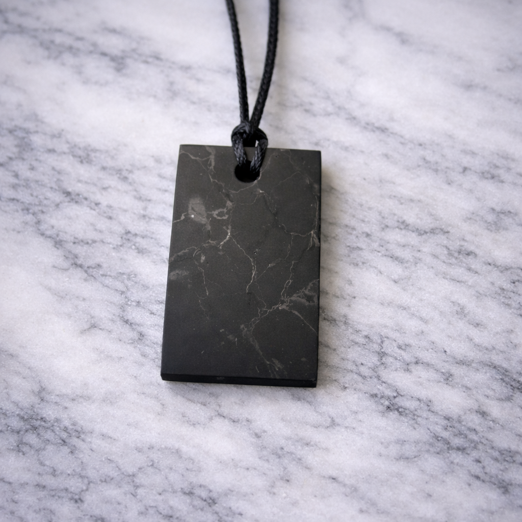 Rectangular shungite pendant on a black cord resting on a white marble surface, showing natural silver mineral flecks and unique stone markings.