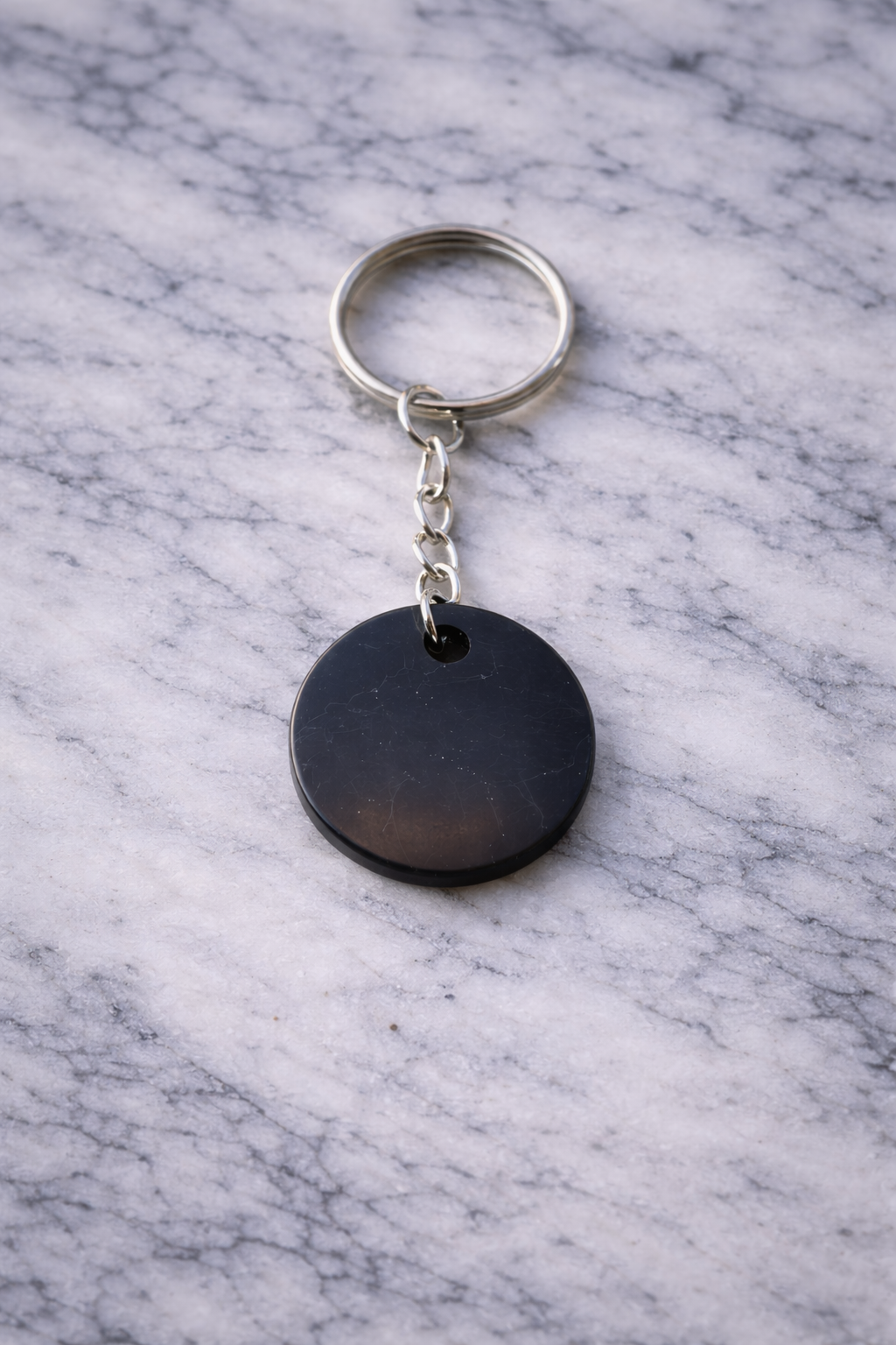 Round polished shungite keychain with silver keyring displayed on a white marble surface, showing smooth black disc with natural stone markings.