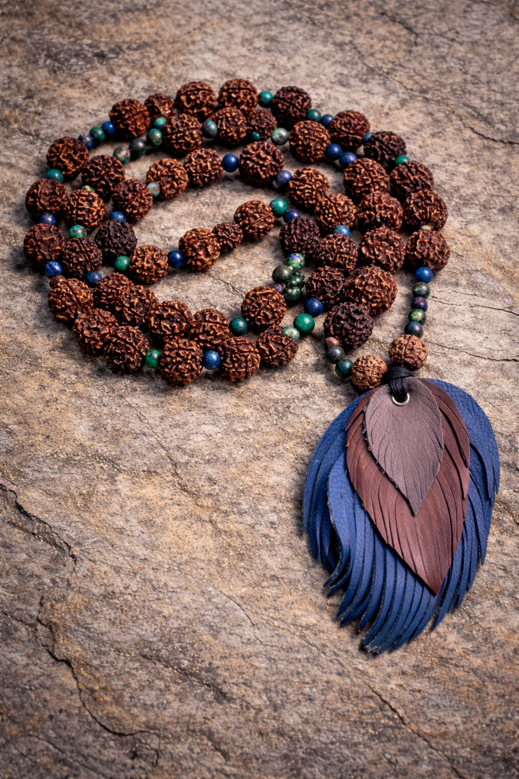 Handcrafted Rudraksha mala with green and blue gemstone accents, coiled on a natural stone surface, finished with a layered brown and blue leather tassel.