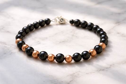 Shungite and copper-coated beaded necklace arranged in a circle on a light marble surface with soft natural lighting and gentle shadows.